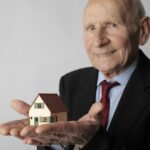 Elderly man in business attire holding a small house model, symbolizing real estate investment.