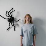 Woman stands with a large artificial spider on wall, portraying fear or anxiety.